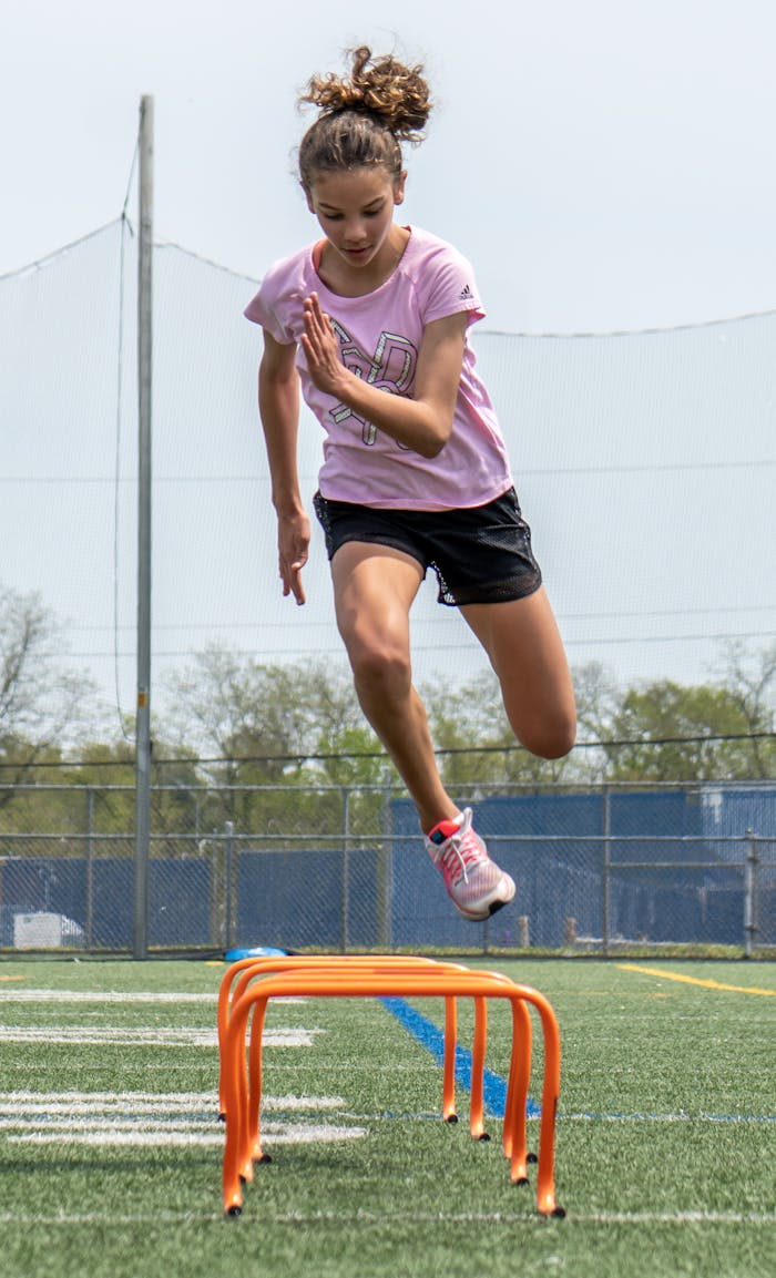 A young woman athlete jumps hurdles on a soccer field showcasing fitness and agility.